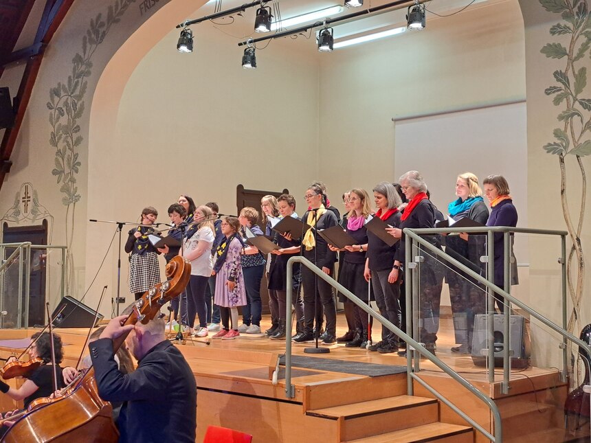Der Schüler- und Lehrerchor steht auf der Bühne und singt. Davor steht ein Orchester.