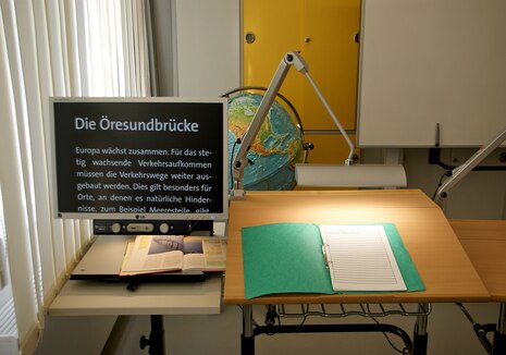 Ein eingerichteter Arbeitsplatz mit einem Bildschirm, auf dem ein vergrößerter Text angezeigt wird. Auf dem Tisch daneben liegt ein aufgeschlagener Hefter.