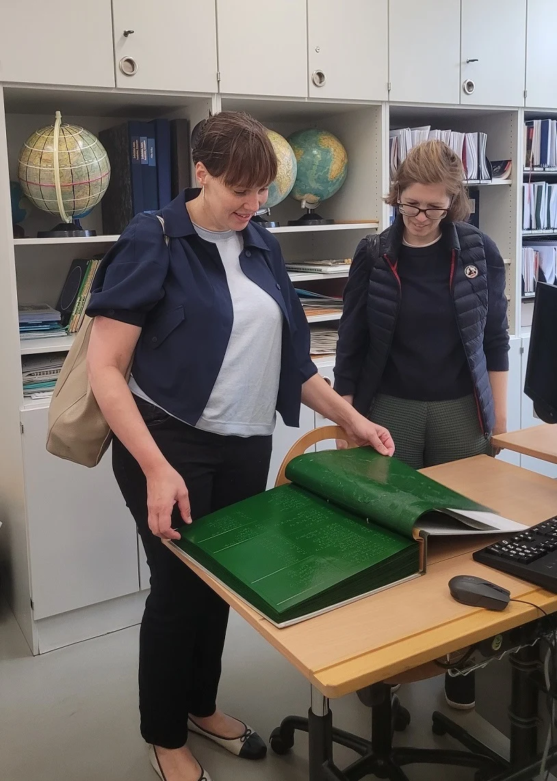 Zwei Frauen stehen an einem Tisch. Darauf liegt ein großes Buch mit Brailleschrift, das sie sich anschauen.