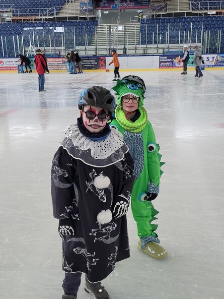 Zwei Jungen, die als Clown und Drache verkleidet sind stehen auf der Eisfläche und schauen in die Kamera. Hinter ihnen sind weitere Kinder zu sehen, die sich auf dem Eis bewegen.