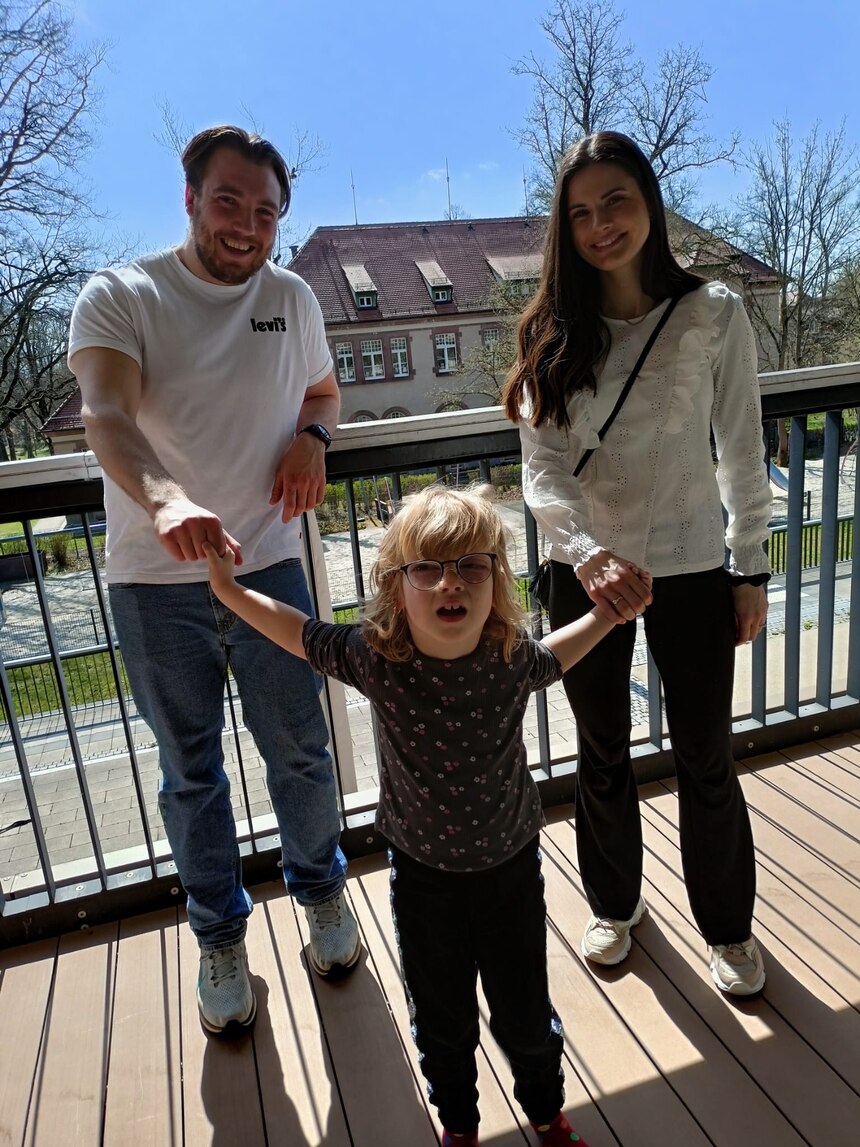 Ein Mann und eine Frau stehen lächelnd auf einer Terrasse und halten eine Schülerin an den Händen. Diese schaut ebenfalls lächenlnd in die Kamera.