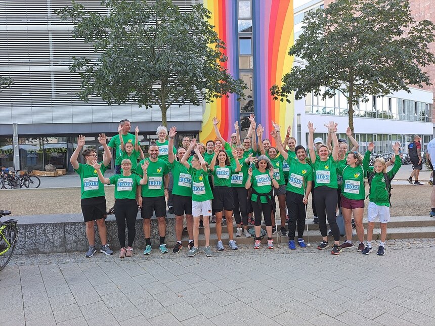 Lehrkräfte, Mitarbeiter und Schüler der Landesschule stehen vor einem bunt bemaltem Gebäude und jubeln mit hochgehaltenden Armen. Sie tragen alle das gleiche T-shirt.