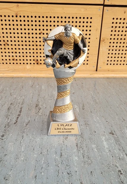 Ein Pokal in den Farben gold und silber. Die Kuppe hat die Form eines Fußballs, in dem ein Fußballspieler mit goldemen Trikot integriert ist.