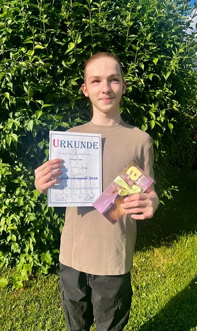 Ein Junge steht vor einem großem Gebüsch und hält eine Urkunde und einen Gutschein in den Händen. Er lächelt stolz in die Kamera.