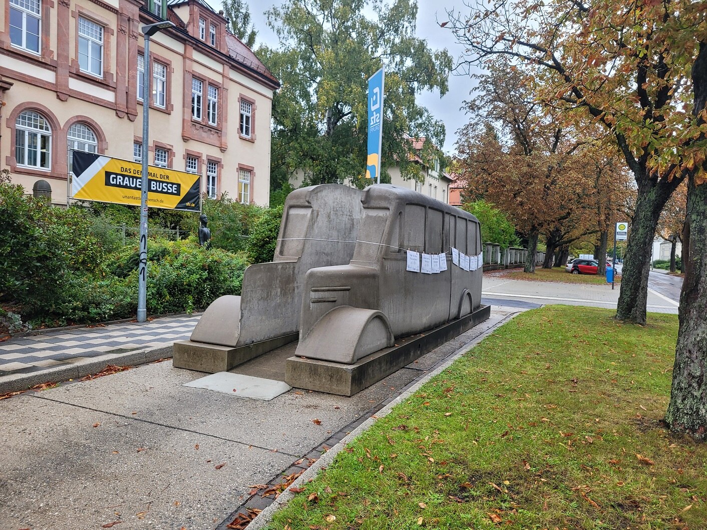 Vor einem Gebäude steht ein Mahnmal aus Beton, in Form eines Busses. An diesem sind an der Seite mehrere Zettel an einer Leine angebracht.