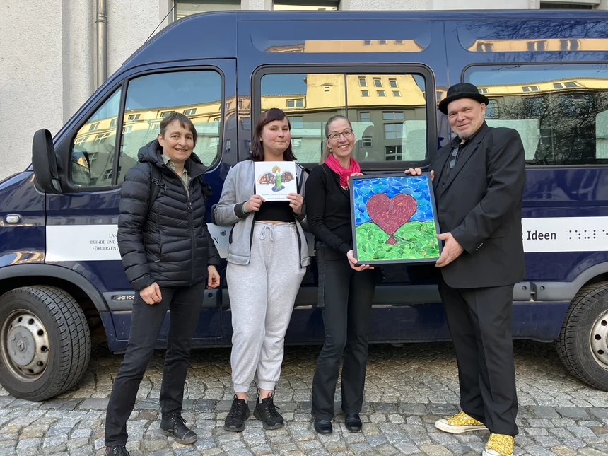 Vor unserem Schulbus stehen ein Künstler, Frau Kauer, Samantha und Frau Stephani. Der Künstler überreicht ein Kunstwerk, auf dem ein Herz aus Mosaiksteinen zu sehen ist.