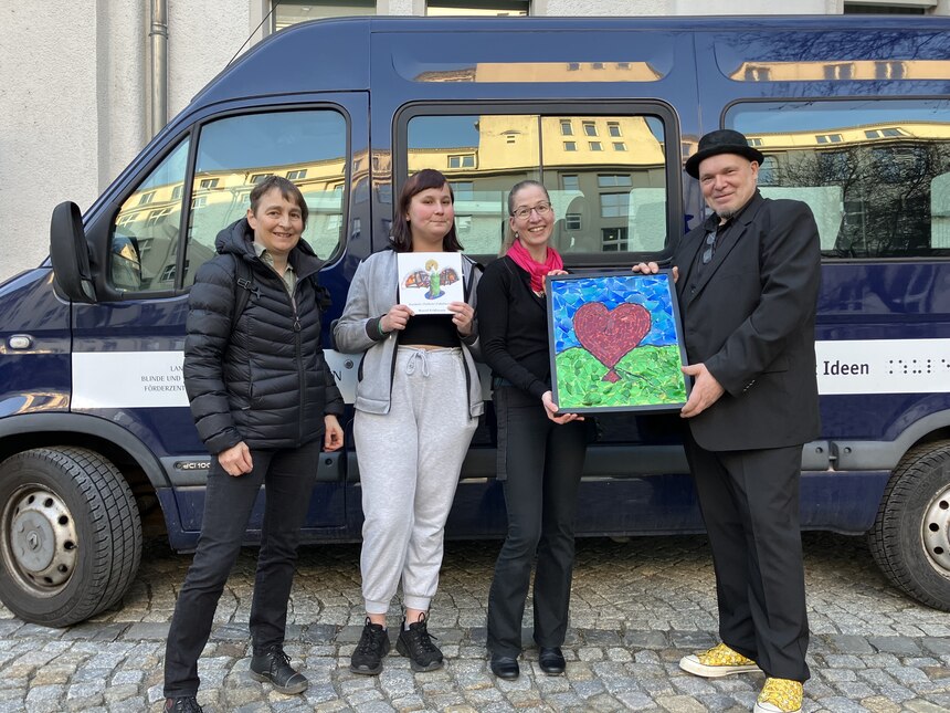 Vor unserem Schulbus stehen ein Künstler, Frau Kauer, Samantha und Frau Stephani. Der Künstler überreicht ein Kunstwerk, auf dem ein Herz aus Mosaiksteinen zu sehen ist.