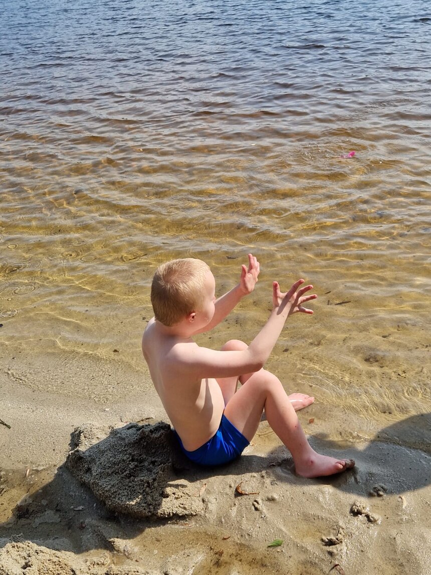 Ein Junge sitzt im Sand am See und spielt mit dem Wasser.