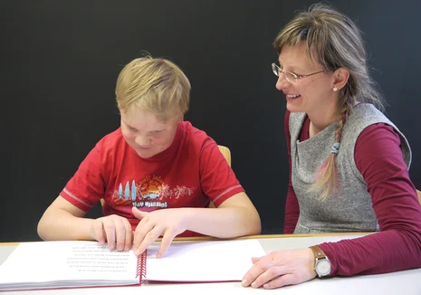 Ein Schüler liest in einem Buch mit Brailleschrift. Daneben sitzt eine Lehrerin.