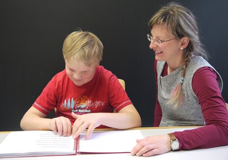 Ein Schüler liest in einem Buch mit Brailleschrift. Daneben sitzt eine Lehrerin.