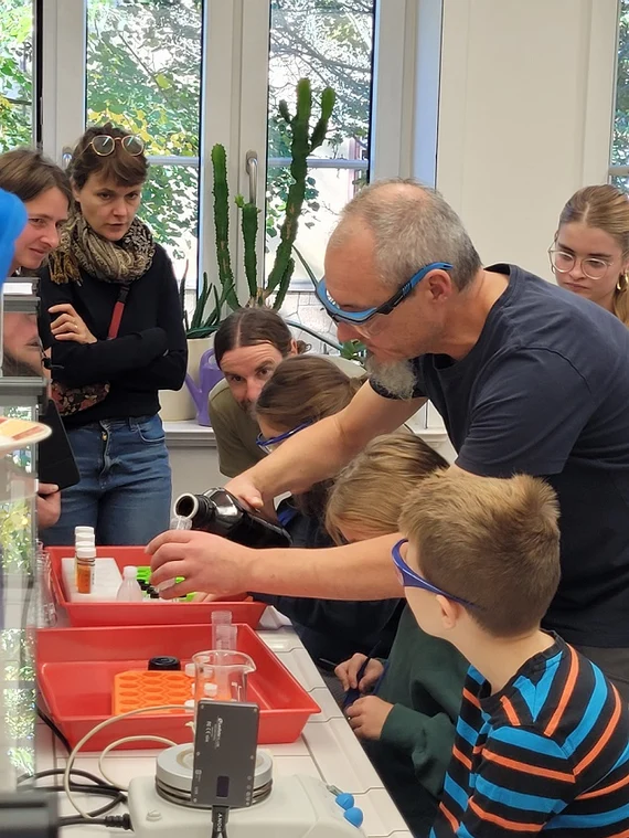 Eine Gruppe vom Erwachsenen und Kindern stehen in einem Chemielabor um einen Experimentiertisch. Ein Mann schüttet eine Flüssigkeit in ein Gefäß. Alle beobachten gespannt, was passiert.