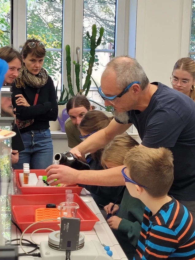 Eine Gruppe vom Erwachsenen und Kindern stehen in einem Chemielabor um einen Experimentiertisch. Ein Mann schüttet eine Flüssigkeit in ein Gefäß. Alle beobachten gespannt, was passiert.