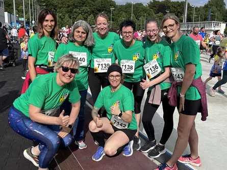 Eine Gruppe von 8 Frauen in Sportkleidung und grünen T-Shirts steht auf einem Sportplatz und lächeln in die Kamera. Im Hintergrund sind viele Menschen zu sehen, die rennen.
