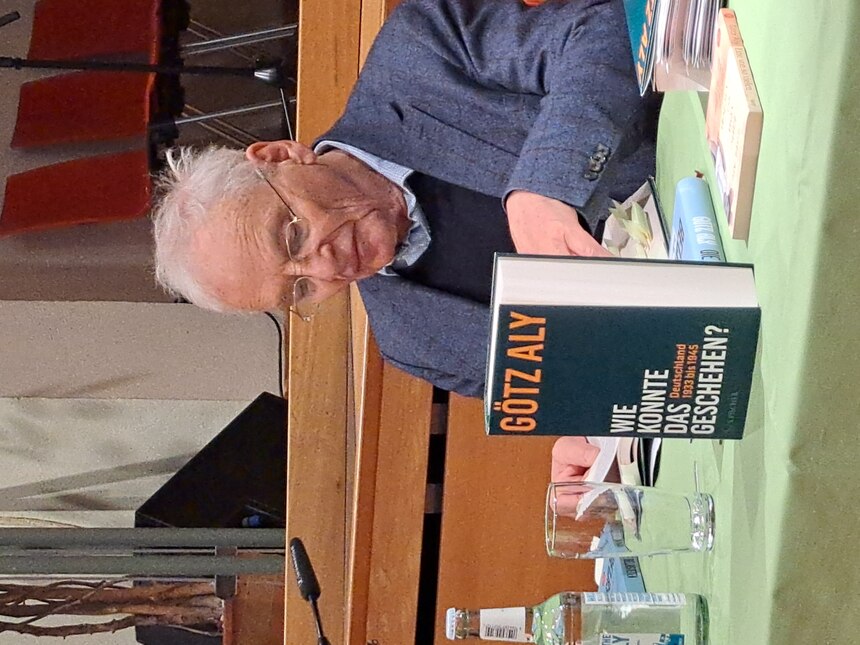 Ein Mann sitz an einem Tisch und blättert in einem Buch. Vor ihm steht ein Buch mit dem Titel "Götz Aly - Wie konnte das geschehen?"