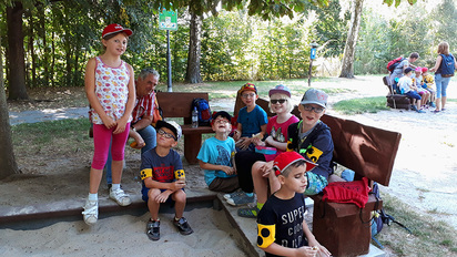 Sieben Schüler der Klasse 2a genießen mit einem Lehrer ihre Pause auf dem Abenteuerspielplatz in Stollberg. Im Hintergrund sitzen weitere Kinder auf einer Bank mit zwei Betreuerinnen.