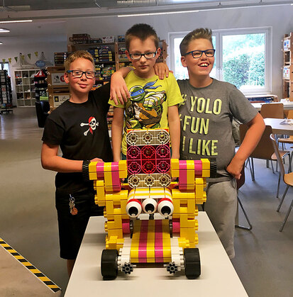 Drei Jungen aus der Klasse 5 stehen im Spielemuseum stolz hinter einem Tisch, auf dem ihr gemeinsam gebautes ziemlich großes „Modellauto“ steht.