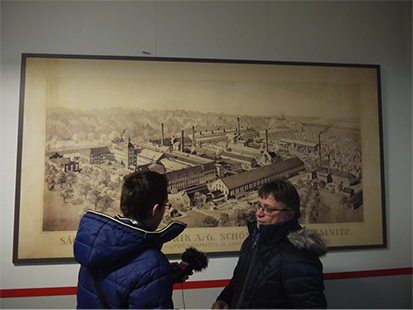 Phillip interviewt die Gaestefuehrerin mit einem Aufnahmegeraet. Im Hintergrund haengt eine historische Zeichnung der Schoenherr-Fabrik an der Wand