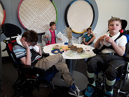 Vier Schüler, zwei davon im Rollstuhl, sitzen um einen Tisch herum auf dem frisches aufgeschnittenes Brot, Brötchen und Behälter mit Aufstrich stehen.