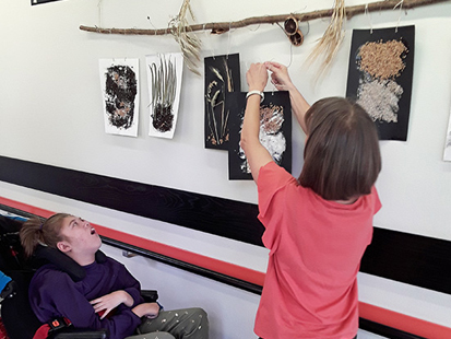 Frau Kaufmann hängt ein mit Körnern gestaltetes Blatt an eine Befestigung und eine Schülerin im Rollstuhl steht daneben. Sie betrachtet die sechs aufgehängten und selbstgestalteten Bilder.