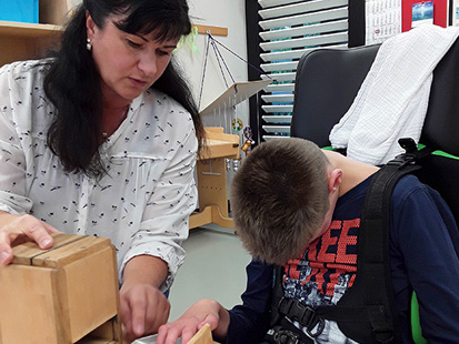 Gemeinsam mit Frau Müller ertastet der Schüler im Rollstuhl das gemahlene Korn, was man in einem kleinen herausgezogenen Schubfach einer kleinen Holzmühle erfühlen kann.