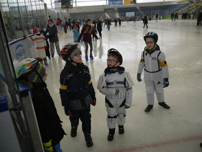 Viele Schüler fahren auf dem Eis mit Schlittschuhen und drei Grundschüler im Vordergrund probieren es erst einmal mit ihren Straßenschuhen.