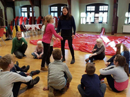 Die kleinen Schauspieler sitzen auf dem Boden im Kreis. Eine Schülerin steht mit Frau Düber in der Mitte und führt besprochene Bewegungen durch.