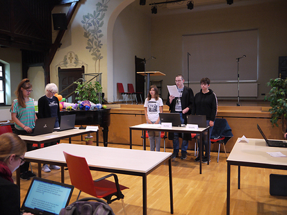 In einem großen Saal stehen fünf Schüler vor ihren Tischen und führen mit verteilten Rollen eine Gerichtsverhandlung durch.