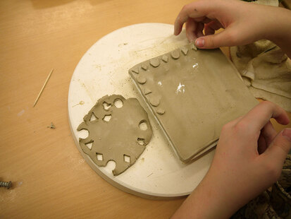 Hier sieht man einen getöpferten quadratischen Teller. Eine Schülerin verziert den Rand des Tellers mit kleinen Dreiecken, Vierecken und Kreisen aus Ton.
