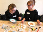 Zwei Schülerinnen sitzen am Tisch und bemalen jeweils einen Porzellanteller mit verschiedenen Farben und Mustern.