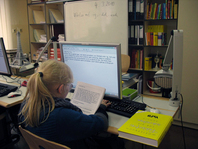 Eine sehbehinderte Schülerin aus der 8. Klasse sitzt im Klassenraum vor ihrem Schülertisch mit Computer. Sie liest im Großdrucklehrbuch und schreibt den Antworttext auf der Tastatur. Die Schülerin arbeitet mit einer Vergrößerungssoftware.