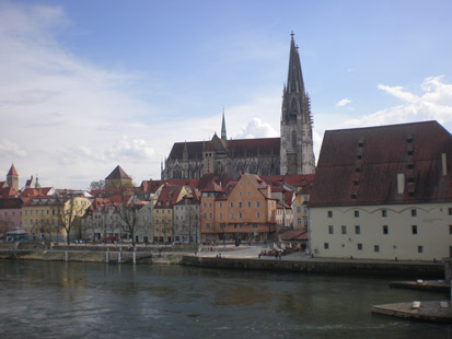 Das Bild zeigt eine historische Häuserzeile von Regensburg am Ufer der Donau. Im Hintergrund ist der Regensburger Dom zu sehen.