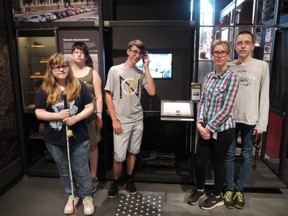 Die Schüler des Neigungskurses Geschichte stehen im Industriemuseum vor einem ihrer Ausstellungsstücke. Max hält den Kopfhörer in der Hand, um die Audiodatei anzuhören. Auf einem Bildschirm kann man Fotos des Projekts anschauen.