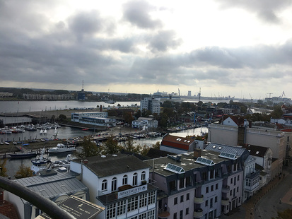 Das Bild zeigt die Aussicht vom Warnemünder Leuchtturm. Es sind der Hafen mit Schiffen und verschiedene Häuser zu sehen.