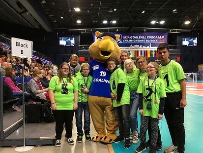 Das Bild zeigt alle Teilnehmer der Reise gemeinsam mit dem Maskottchen Goalino am Spielfeldrand in der StadtHalle Rostock.