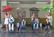 Die vier Schüler sitzen im Vordergrund auf dem Stuhl oder im Rollstuhl. Dahinter stehen Pädagoginnen, die mit einem aufgespannten Regenschirm über den Köpfen einen Liedtext spielerisch umsetzen