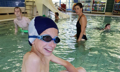 Julian sitzt am Beckenrand vom Nichtschwimmerbecken der Schwimmhalle und lacht verschmitzt in die Kamera.