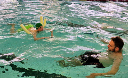 Alrik schwimmt mit einer Nudel unter den Oberarmen im tiefen Becken dem Trainer hinterher.