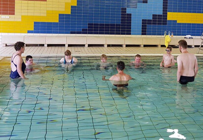 Alle Schüler stehen im Wasser am Beckenrand und hören aufmerksam den Erläuterungen der Schwimmtrainer zu.