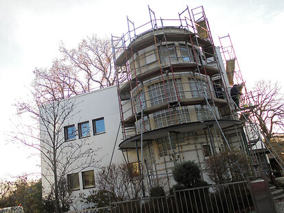 Das Bild zeigt die Vorderansicht des Gebäudes, das momentan saniert wird. Markant ist hier das runde Treppenhaus in Glasbausteinbauweise.