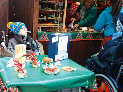 Nils Ebermann sitzt vor dem Weihnachtsstand an einem kleinen Tisch und wirbt für unsere Keramik. Im Hintergrund sieht man Leonie und Frau Bach beim Einpacken einer Keramik.