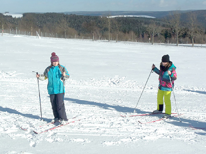 Zwei Maedchen fahren mit Langlaufskiern auf einer verschneiten Wiese.