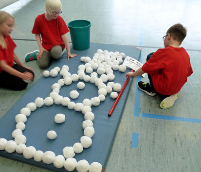 Drei Schüler sitzen um eine Turnmatte herum auf der mit weißen Bällen ein großes Schneemannbild zusammengelegt ist.