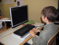 Ein Schüler aus der 8. Klasse sitzt vor dem Schülertisch mit Computer und bearbeitet Aufgaben aus dem Blindenlehrbuch. Das Lehrbuch liegt bei ihm auf den Knien. Die Hände befinden sich auf der Braillzeile und ertasten die angezeigte Blindenschrift. 
