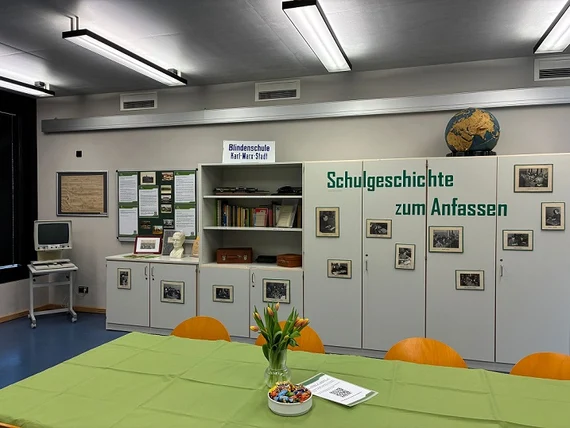 Das Bild zeigt den Einblick in das Museum. An der Wand stehen mehrere Schränke mit der Aufschrift „Schulgeschichte zum Anfassen“. Zudem sind historische Fotografien und Texte an den Schränken angebracht. Im Regal stehen Fachliteratur und Punktschriftmasch