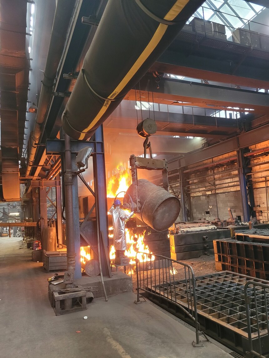 In einer Halle der Gießerei schüttet ein Mann mit Schutzanzug flüssiges Eisenerz aus einem Behälter in eine Form. Überall sind Flammen zu sehen.