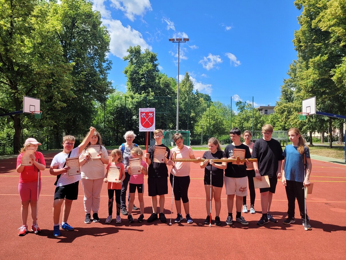 Eine Gruppe von Schülerinenn und Schülern steht auf dem Sportplatz. Jeder hält eine Urkunde für den ersten Platz in der Hand. Einige halten zusätzlich gemeinsam eine großes Schwert aus Holz.