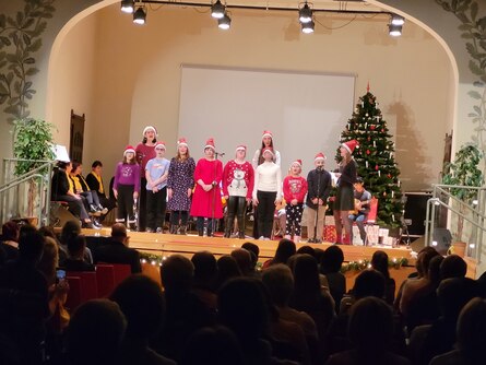 Eine Gruppe von Kindern steht auf einer weihnachtlich geschmückten Bühne und singt.Mit ihnen singen drei Erwachsene. Alle tragen Weihanchtsmützen auf dem Kopf. Vor der Bühne sitzen viele Zuschauer.