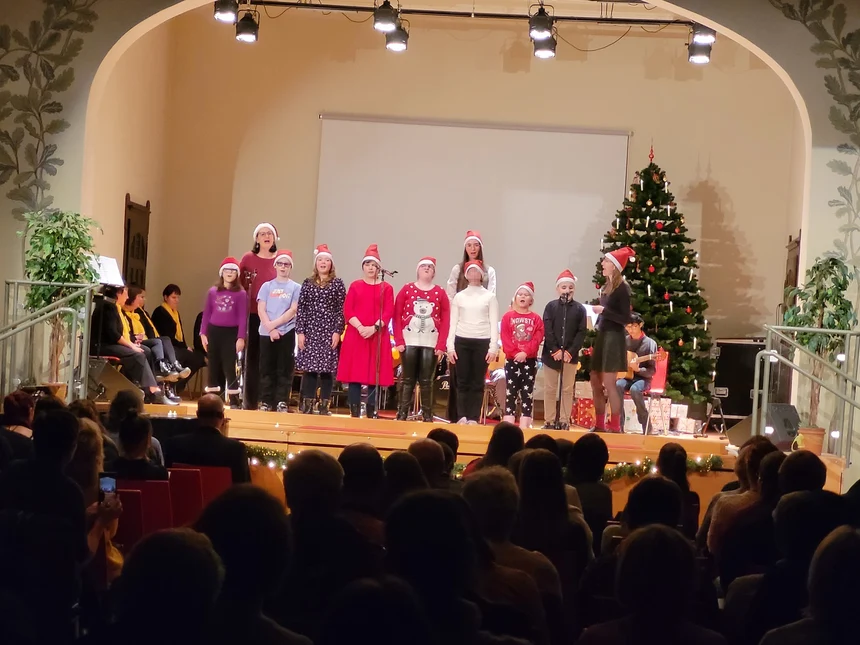 Eine Gruppe von Kindern steht auf einer weihnachtlich geschmückten Bühne und singt.Mit ihnen singen drei Erwachsene. Alle tragen Weihanchtsmützen auf dem Kopf. Vor der Bühne sitzen viele Zuschauer.