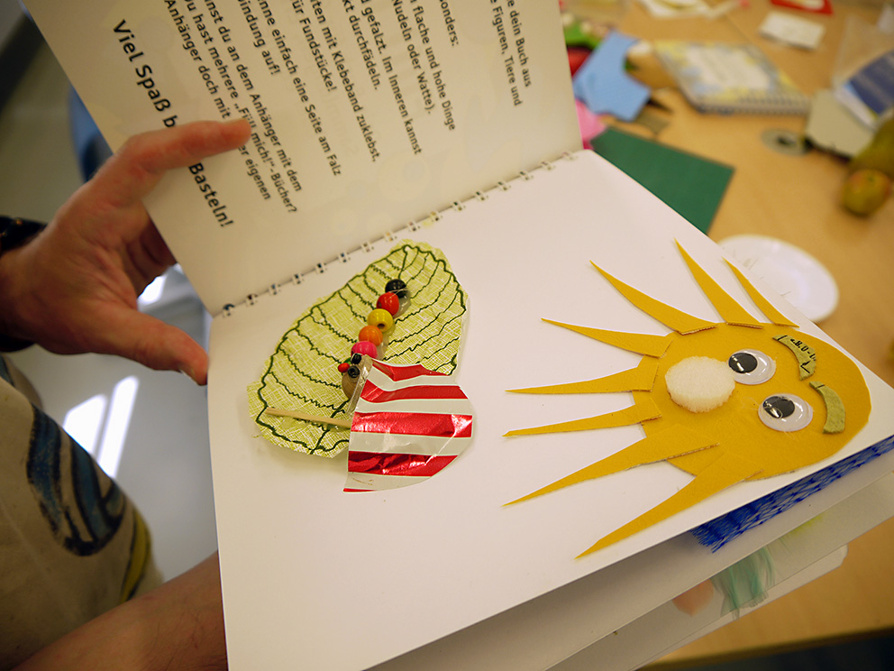Eine Person hält ein aufgeschlagenes Tastbuch in der Hand. Dort wurden eine Sonne und die Raupe Nimmersatt auf einem Laubblatt taktil dargestellt, welche unter einem Sonnenschirm Schatten sucht.