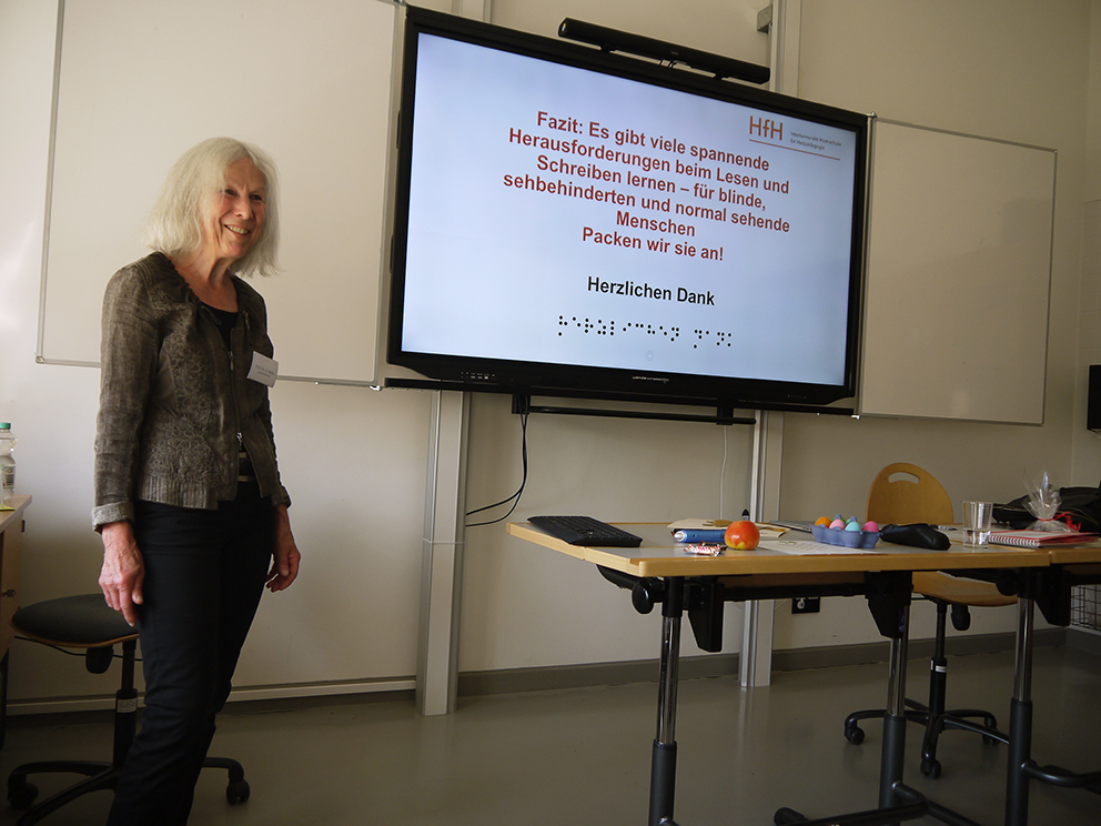 Frau Prof. Dr. Hofer steht lächelnd vor einer digitalen Tafel, auf der das Fazit ihres Vortrags („Es gibt viele spannende Herausforderungen beim Lesen und Schreiben lernen – für blinde, sehbehinderte und normal sehende Menschen. Packen wir sie an!) geschrieben steht.